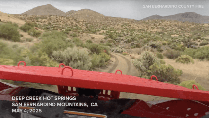 View from the back of a rescue vehicle driving along a rugged dirt trail through desert terrain surrounded by low shrubs and distant mountains. Overcast skies loom above. Bold white text at the bottom reads: “DEEP CREEK HOT SPRINGS, SAN BERNARDINO MOUNTAINS, CA, MAY 4, 2025.” “SAN BERNARDINO COUNTY FIRE” is also visible in the top right corner.